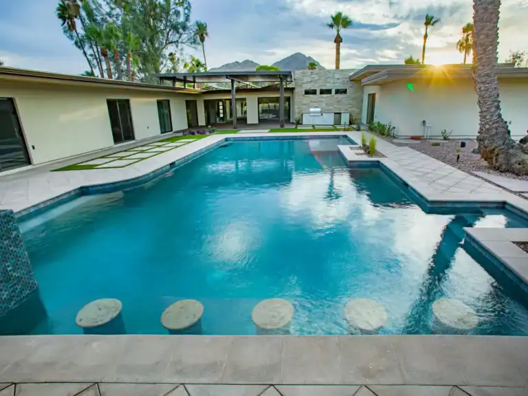 Modern desert backyard with large swimming pool, swim-up bar stools, outdoor kitchen, and mountain views at sunset.