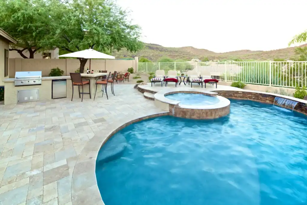 Desert backyard with outdoor kitchen, grill, bar seating, swimming pool, hot tub, and stone waterfall.