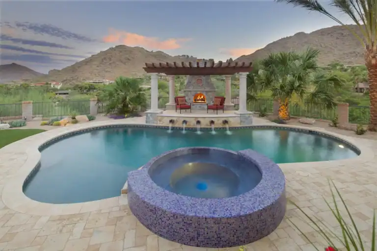 Luxury backyard pool and spa with a cozy outdoor fireplace, elegant pergola, and stunning mountain views at sunset.