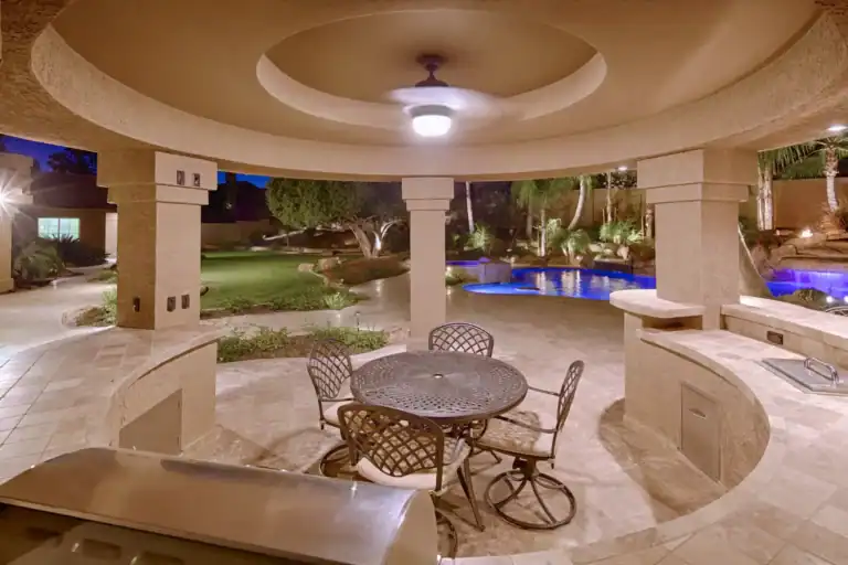 Luxury outdoor kitchen under a curved patio, with grill, dining set, and illuminated backyard pool at night.