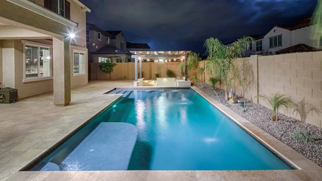 An exquisite, tranquil backyard pool illuminated at night, featuring shimmering azure water and a lavish patio area