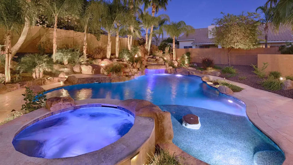 Lavish backyard oasis at twilight: an opulent illuminated pool, tranquil hot tub, and dazzling waterfall set among vibrant, uplit palm trees