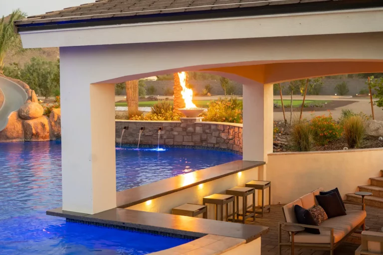 Exquisite poolside retreat featuring a lavish covered bar, elegant seating overlooking a vibrant blue pool with water features, and a dramatic fire bowl backdrop