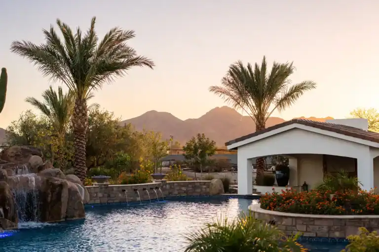 A sumptuous Arizona pool oasis bathed in the radiant light of sunset, featuring a cascading rock waterfall, elegant water spouts, lush palm trees, and a covered seating area
