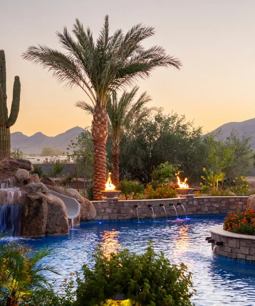Exquisite desert oasis pool with cascading rock waterfall, winding slide, and elegant fire bowls glowing at dusk, surrounded by lush palms and a majestic saguaro cactus